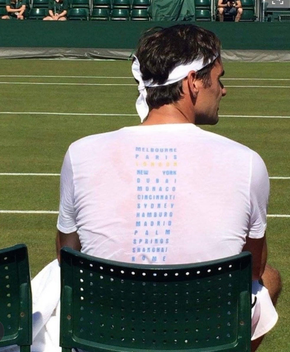 A player seated courtside, seen from behind, headband on, a list of cities printed down the shirt's back
